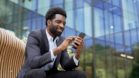 Smiling african american businessman in formal suit using mobile phone while sitting on a bench on street near modern office building. Happy worker checks messages, chats online, browses web. Close up - Powered by Shutterstock - Get 15% off with code: PIKWIZARD15