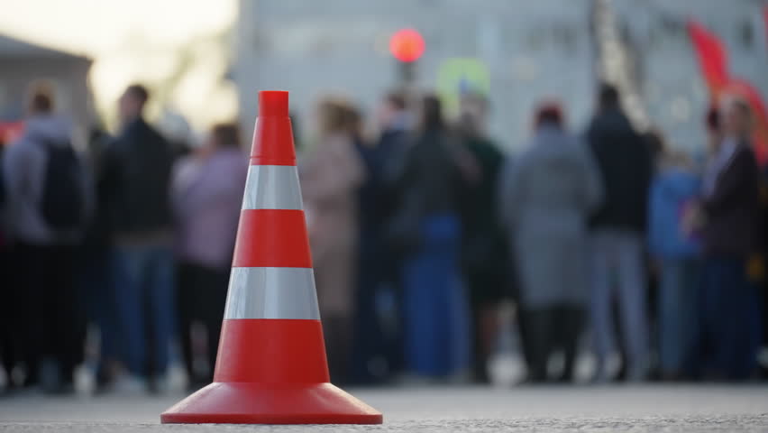 Close-up of an orange traffic cone placed on a paved street with a blurred crowd of people in the background, suggesting a public gathering or event scene.