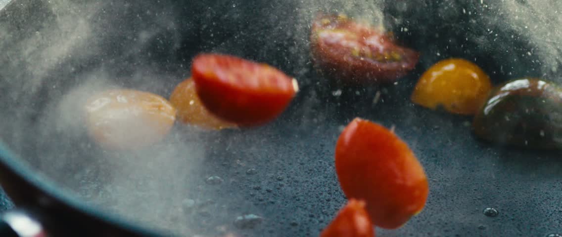Vibrant cherry tomatoes fall into a hot pan and sizzle in super slow motion, releasing a cloud of steam. A dramatic shot for recipe videos or food ads.