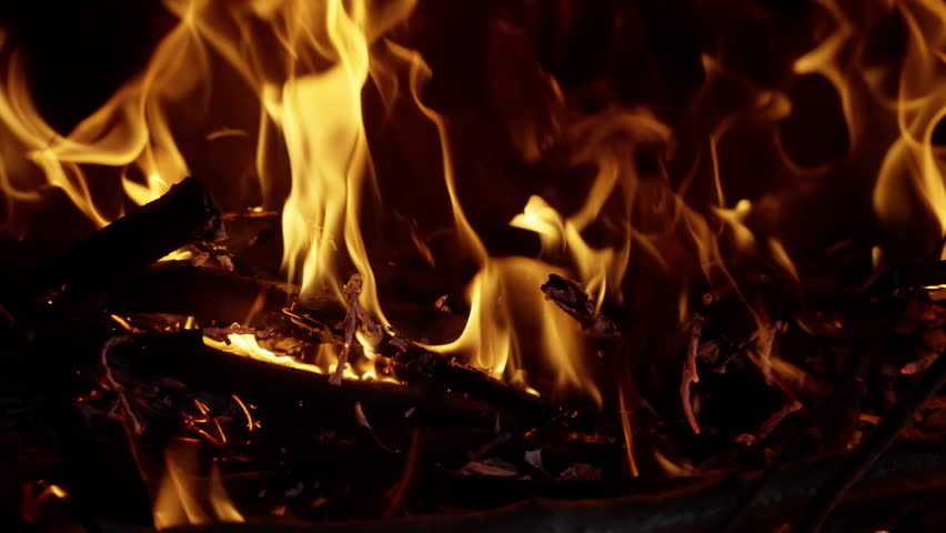 Close-up detailed view of bright orange flames consuming dry wood and kindling, displaying the textures and intensity of a controlled fire burning in darkness.