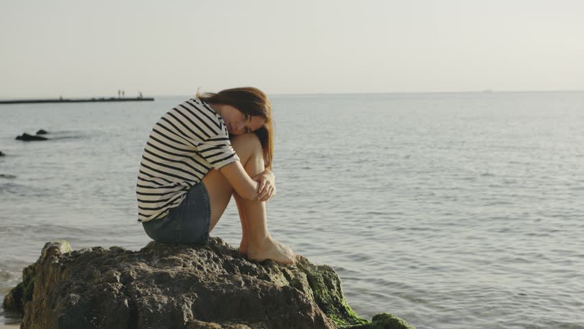 Contemplative moment by the seaside during sunset, featuring a woman sitting on a rock by the calm ocean waves