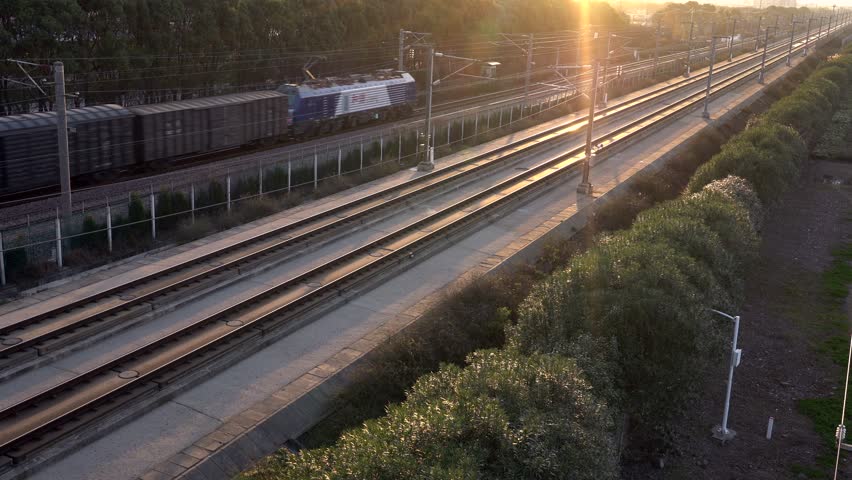 A freight train travels along the railroad tracks bordered by lush greenery and modern infrastructure as the sun sets, casting a warm glow over the scene.