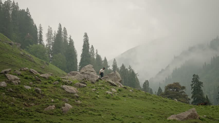Minimal wide shot of Indian man standing on a rock along a mountain trail, small figure in vast natural landscape, 4k video