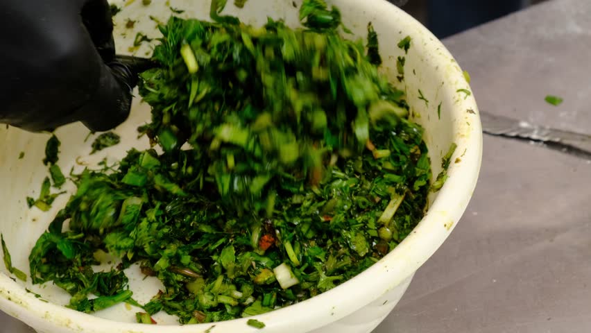 A chef stirs chopped greens in a bowl with a large spoon preparing filling for flatbread made with many mountain herbs Vegetarian version of Jingyalov Hats containing dozens of different herbs