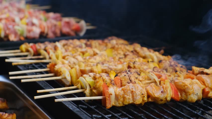 A grill chef uses metal tongs to turn juicy barbecue made of white poultry meat on a hot outdoor grill at an open-air restaurant. Grilled low-fat meat served with vegetable pieces for healthy eating.