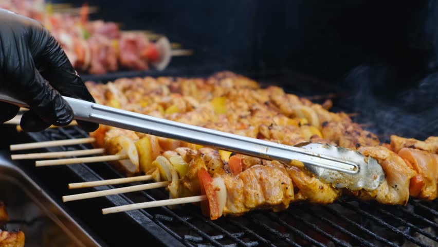 A grill chef uses metal tongs to turn juicy barbecue made of white poultry meat on a hot outdoor grill at an open-air restaurant. Grilled low-fat meat served with vegetable pieces for healthy eating.