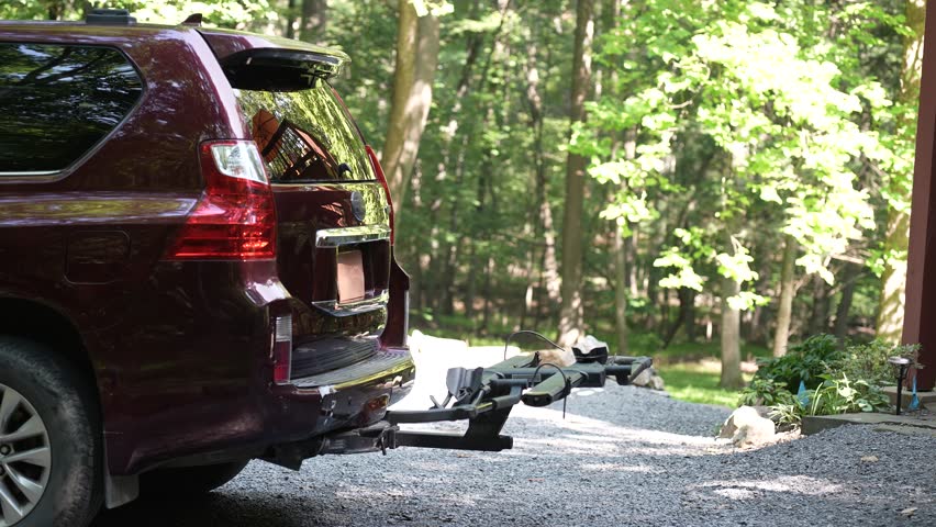 A person is securely placing a bike onto a hitch-mounted rack on the back of a car in a scenic wooded location, preparing for an outdoor biking adventure.