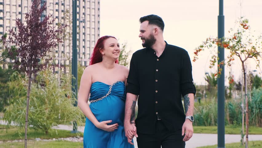 A pregnant woman in a blue dress and her partner walk hand in hand through a public urban park. They lovingly look at each other, enjoying the moment of expecting their baby
