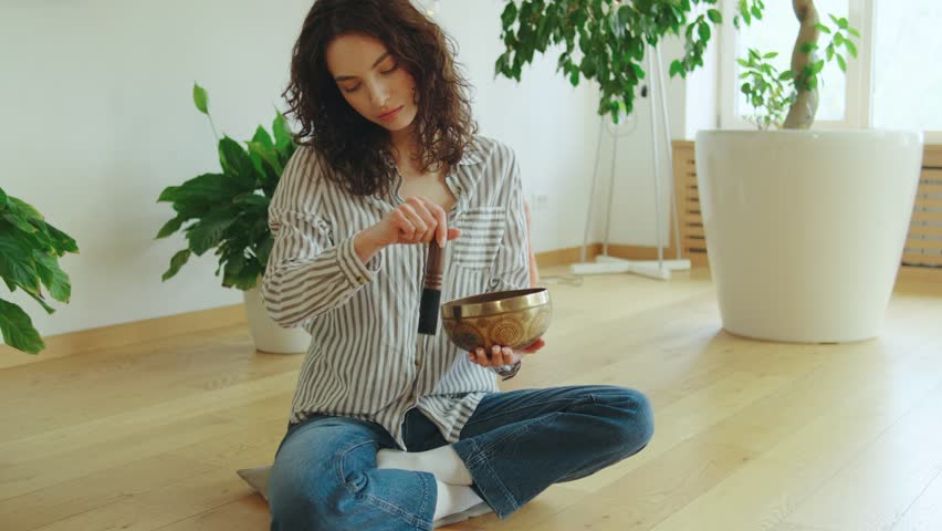 Woman practicing meditation and mindfulness while using a singing bowl in a serene indoor space with greenery