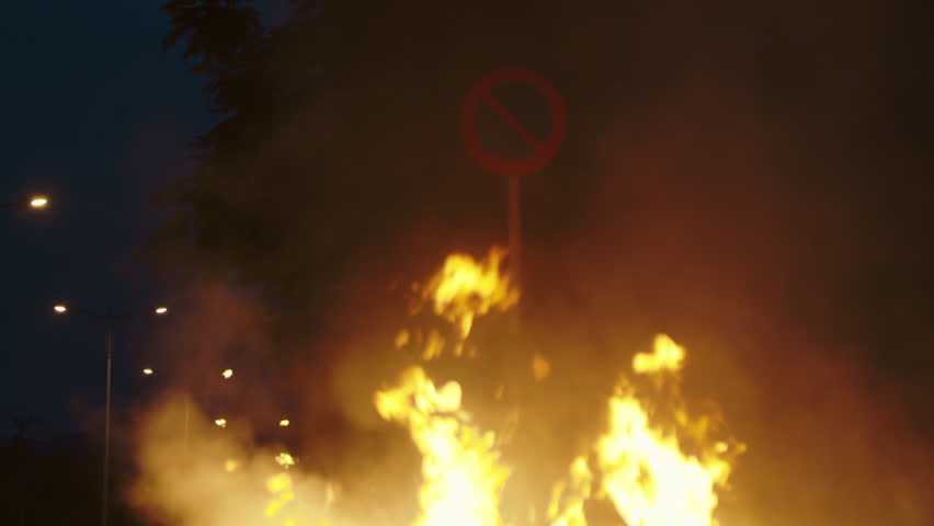 Fire burns on deadly car collision place near road sign at night. Flame destroys broken automobile carcass in evening urban environment. Traffic accident dangerous situation