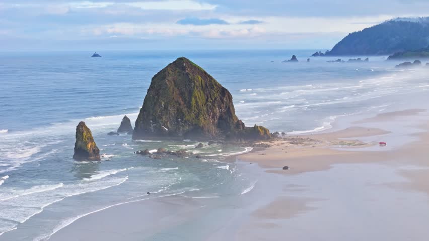Aerial telephoto orbit of Haystack Rock at Cannon Beach, Oregon on a foggy morning with waves rolling onto the beach and moody coastal scenery