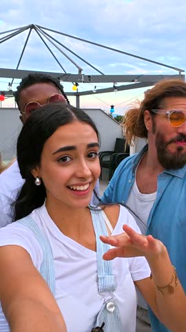 Cheerful multi ethnic friends capturing rooftop party moments, sharing joyful selfies against vibrant urban evening backdrop