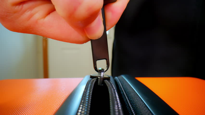 Close up view of fingers holding and zipping suitcase against the background of the white door. Fingers slowly fasten zipping zipper on packed orange luggage baggage suitcase bag