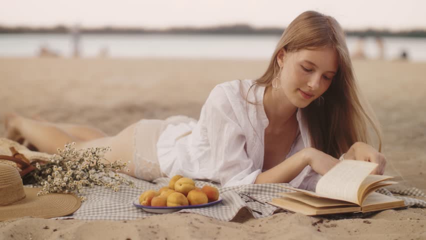 A beautiful blonde woman lies on the sandy beach, enjoying a calm picnic while reading a book. Surrounded by fresh apricots and flowers, this serene summer scene radiates relaxation and tranquility.