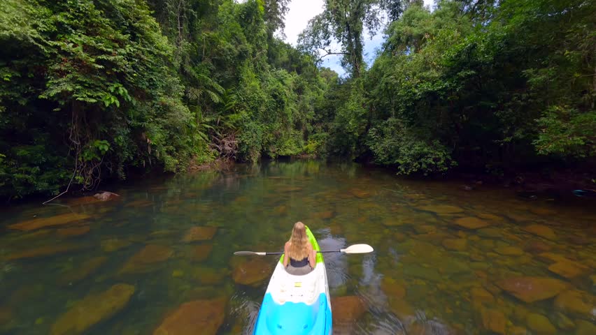 FPV drone shot of woman kayaking on tropical river in lush green jungle in Thailand. Peaceful, cinematic nature scene ideal for travel, ecotourism, and outdoor lifestyle projects.