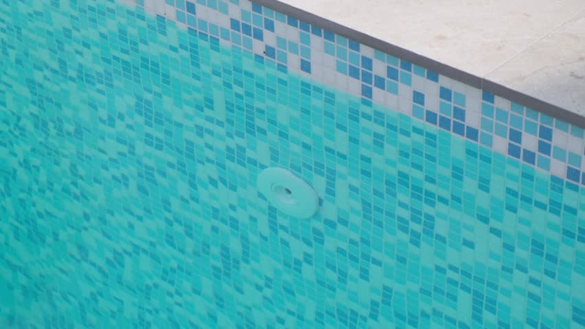 Fragment of a swimming pool wall covered with blue mosaic tiles, featuring a visible water inlet nozzle. Clear water creates ripples that form a pleasant surface pattern
