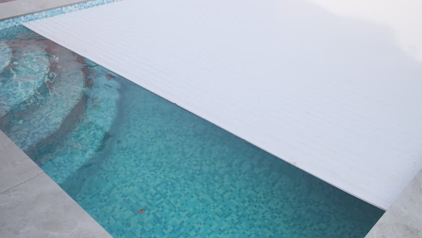 Swimming pool partially covered by an automatic white slatted roller cover. The left side reveals the water, steps, and tiled floor, while the right side is protected
