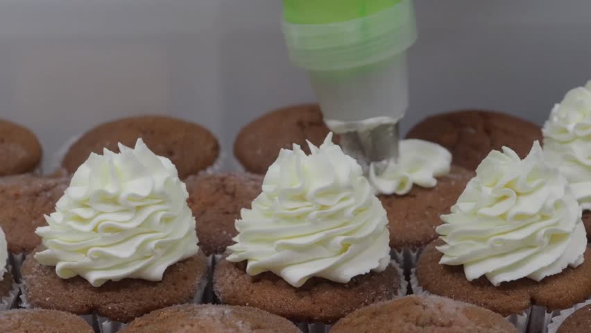 Pastry chef skillfully decorating cupcakes with smooth white cream from a piping bag, creating an exquisite dessert in a bustling professional kitchen filled with delicious aromas slow motion
