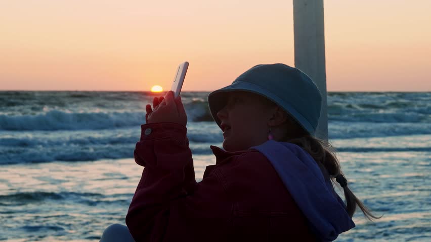 Teenage Girl Filming Sunset by the Sea on Smartphone – Casual Beach Evening