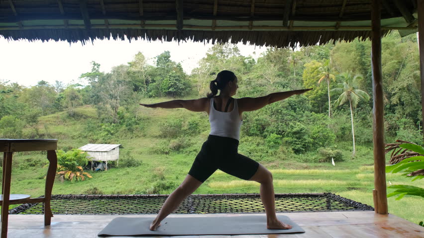 Yoga session unfolds on a serene bamboo hut terrace, as a young woman finds balance amidst lush tropical greenery and rice plantations. Active lifestyle and wellness concept