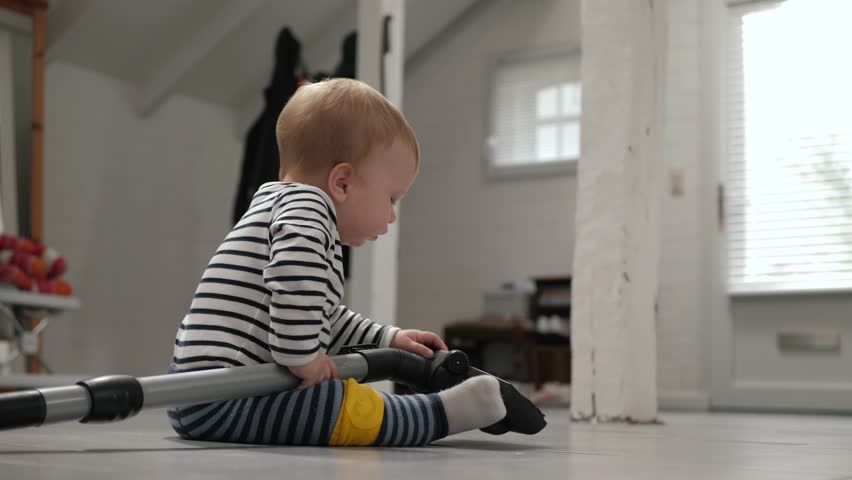 Curious infant in striped clothes exploring vacuum cleaner on heated floor. Development, growth and exploration of home appliances for little boy