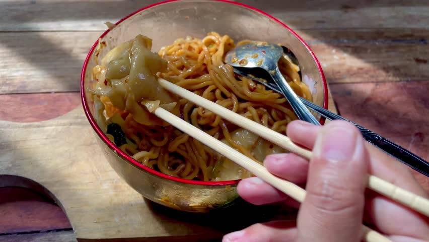 A full bowl of chicken noodles on a wooden cutting board with a wooden table background, typical Indonesian chicken noodles with dumpling toppings, it looks like someone is eating with chopsticks
