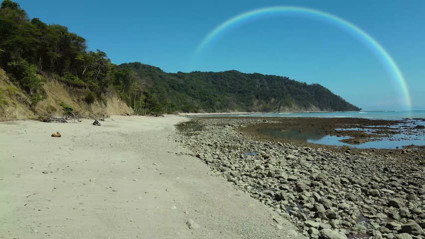 A stunning rainbow arches across the sky after rainfall, captured from a serene aerial perspective above the beach.