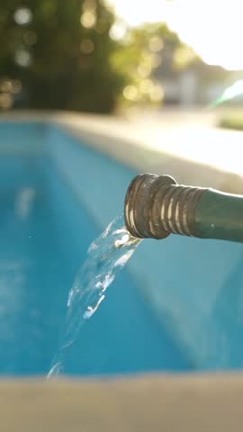 Vertical Close up stream of water flowing from a green hose and filling a clean blue swimming pool in the backyard at sunset. Weekly pool maintenance and cleaning