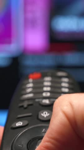 Vertical close-up of a smart TV remote control and man's hand turn on and off, scrolling and choosing content for watch on online video platform. Blurred television screen in the background