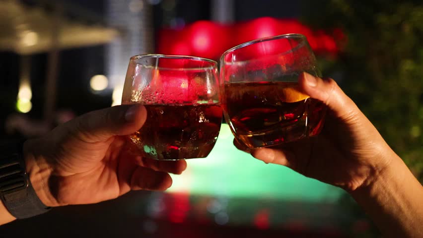 Two people clink whiskey glasses in a celebratory toast beside a brightly lit outdoor pool at night, with warm, festive lighting and shallow depth of field - Powered by Shutterstock - Get 15% off with code: PIKWIZARD15
