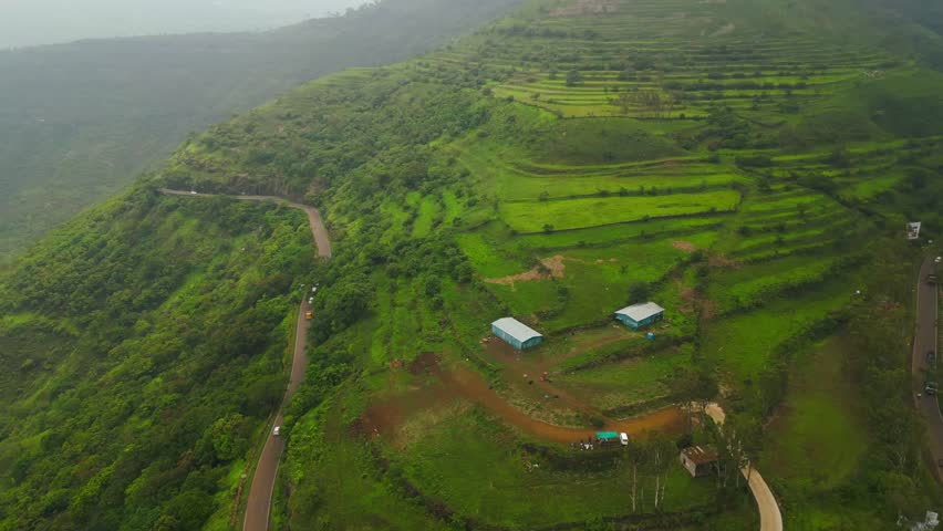An aerial view of a ghat road in Panchgani, Maharashtra, shows vehicles winding through lush green mountains under a dramatic monsoon sky, capturing the season’s vibrant and misty charm 4K