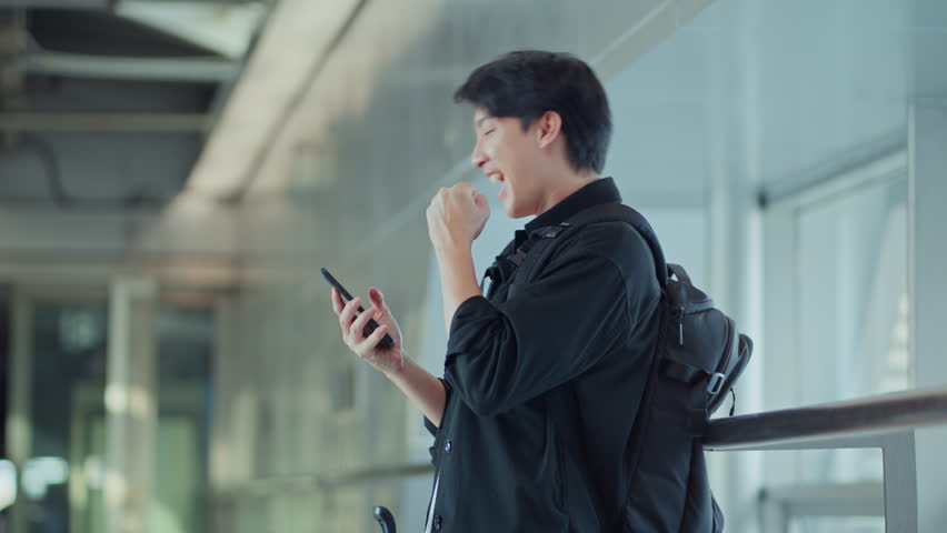 Excited asian male traveler with backpack using smartphone in airport walkway after booking flight on time – happy travel moment, mobile app lifestyle