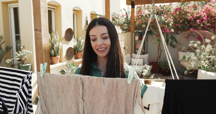 Happy Latin housewife hanging out wet clothes on backyard after laundry, enjoying household work, domestic activity outside, housekeeping on weekend at home, smiling