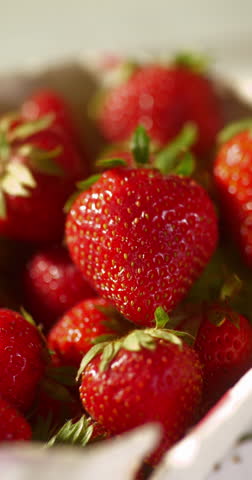 Extreme close-up of a hand gently lifting a ripe strawberry from a full box. The juicy red berries glisten in soft natural light, emphasizing their freshness and vibrant texture.