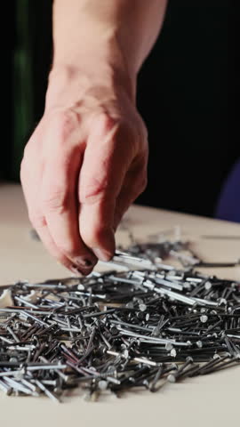 Man actively gathers scattered pile of dark metallic nails consolidating into heap on light surface. Manual process of organizing building materials
