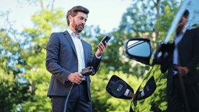 Successful businessman in a sharp suit happily charges electric car outdoors. Modern professional uses smartphone to manage charging, sustainable transportation and smart technology integration. - Powered by Shutterstock - Get 15% off with code: PIKWIZARD15