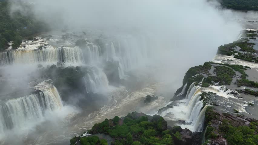 Aerial view of the Iguazu Falls marks border between Brazil and Argentina. Mist rises from the canyon. Water streams plunge in multiple tiers. Forests surround the famous U-shaped Devil