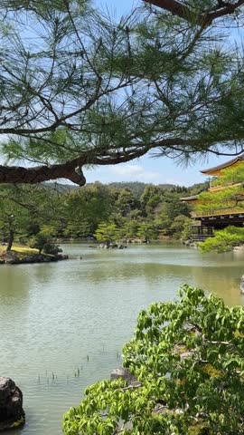 Kinkaku-ji Temple, the Golden Pavilion, in Kyoto, Japan, reflects in a serene pond under a bright, cloud-filled sky. Ancient trees frame the iconic gilded building, a sight of unparalleled beauty.