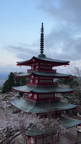 Majestic Mount Fuji, partially veiled in clouds, provides a breathtaking backdrop to a vibrant cherry blossom scene in Japan. A traditional red pagoda adds cultural richness to this tranquil