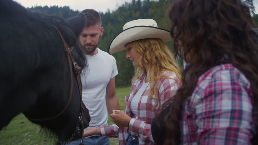 Cheerful young couple visiting a Western-style riding school, connecting with an instructor and a friendly horse as they feed it by hand, medium close up shot.
