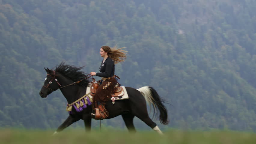 Powerful footage of a skilled cowgirl on horseback, riding a beautiful black horse through a mountain wilderness, slow motion shot.