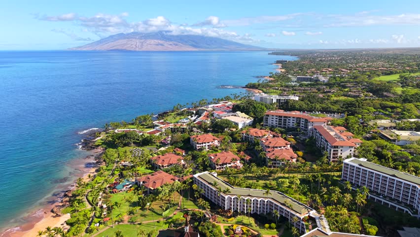 Luxury resorts on tropical coast of Wailea in Maui with distant island, aerial push-in shot