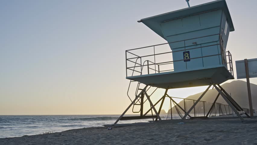 A static shot of a lifeguard tower silhouetted against the setting sun on a quiet California beach, with soft waves rolling in and golden hour light casting long shadows.