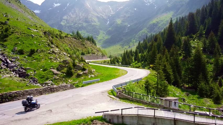 Drone tracks biker riding through Transfăgărășan road in Carpathian Mountains of Romania, surrounded by lush greenery and alpine landscape.