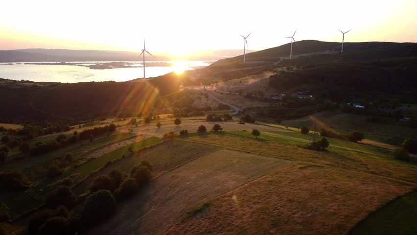 Český Banát-Aerial drone push forward over Romania's rural fields at sunset, wind turbines spin over farmland and hills with lake views.