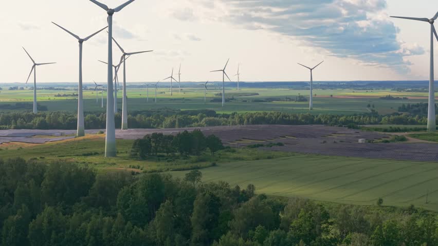A drone view of expansive green rural landscape dotted with tall wind turbines, harnessing renewable energy under a partly cloudy sky at dusk