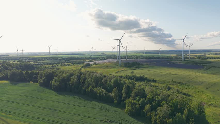 A drone view of green rural landscape dotted with wind turbines, harnessing renewable energy under a partly cloudy sky