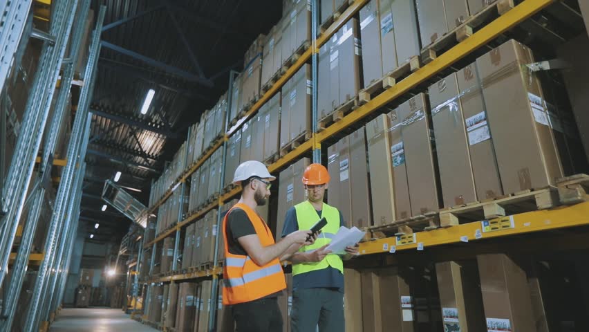 A warehouse worker and a storekeeper work in a large multi-level warehouse, inside. Accounting for goods in a factory finished goods warehouse. Large multi-level warehouse with cardboard boxes on rack