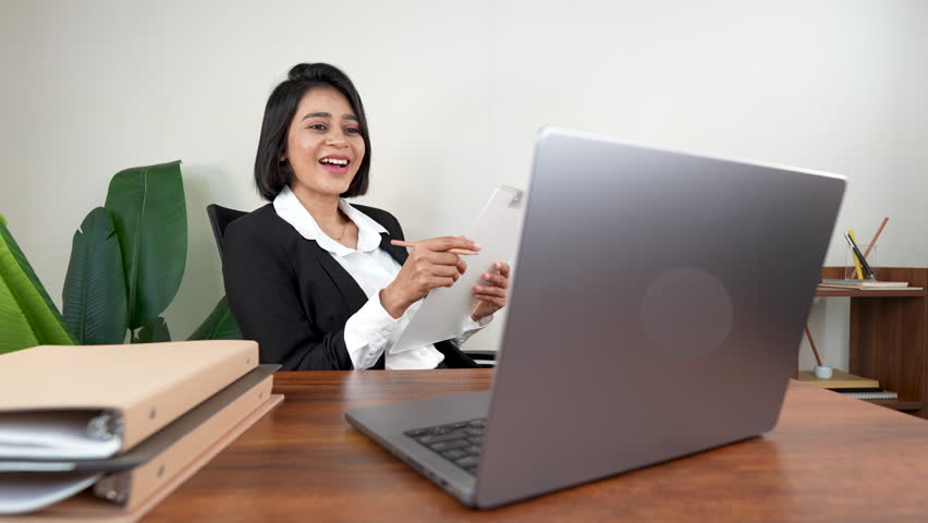 A cheerful woman with short dark hair laughs heartily while looking at her laptop at a wooden desk in an office