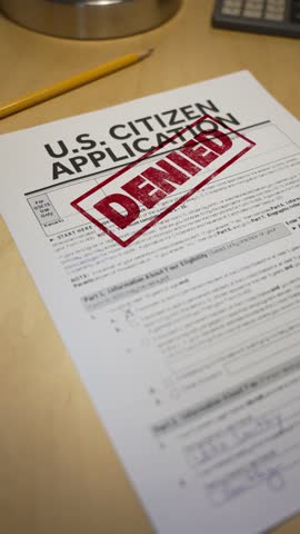 Vertical view of a U.S. citizenship application form with a bold red “DENIED” stamp. A pencil rests nearby, highlighting the rejection of the naturalization request on a wooden desk surface. 14710  	
